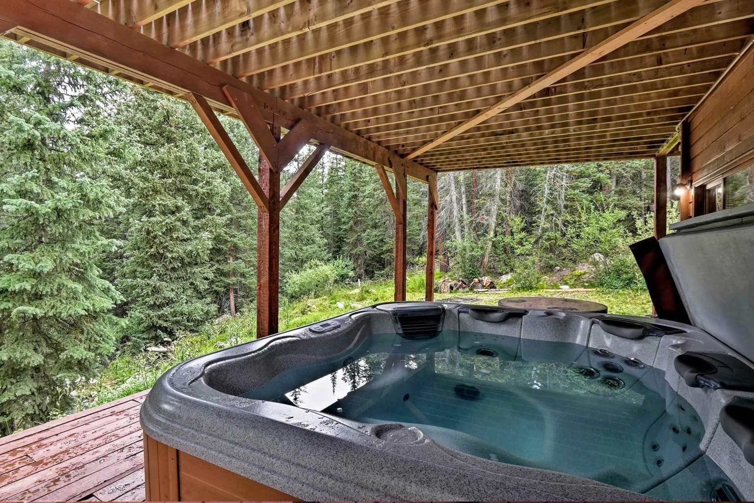 Hot tub on a wooden deck under a pergola, overlooking a dense evergreen forest.