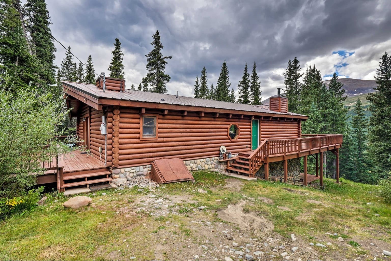 Rustic log cabin with a wooden deck surrounded by pine trees under a cloudy sky.